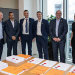 6 people stood around a table with reams of paper laid out